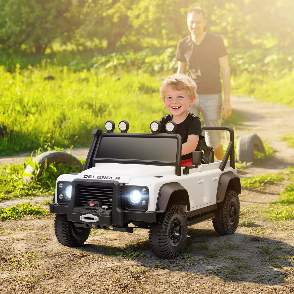 Berniukas vairuoja baltą Land Rover Defender elektromobilį su nuotolinio valdymo pultu šalia