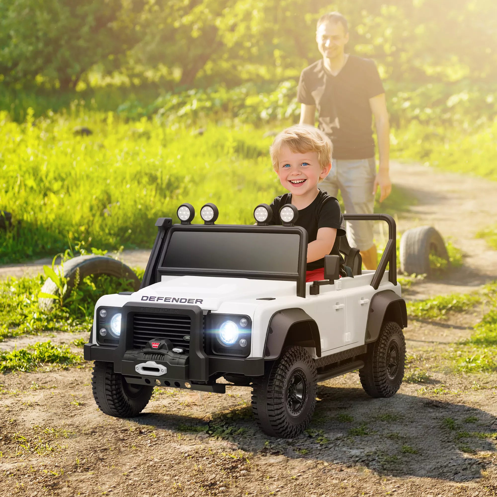 Berniukas vairuoja baltą Land Rover Defender elektromobilį su nuotolinio valdymo pultu šalia