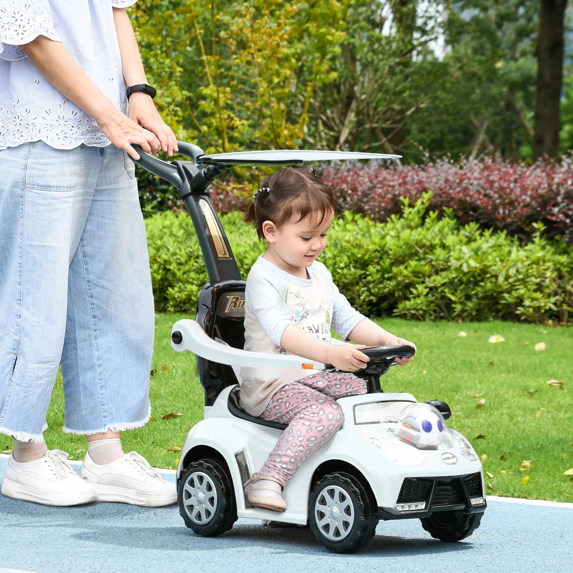 Mergaitė sėdi baltame stumiamuose automobilyje lauke, tėvas stumiamas iš galo rankena