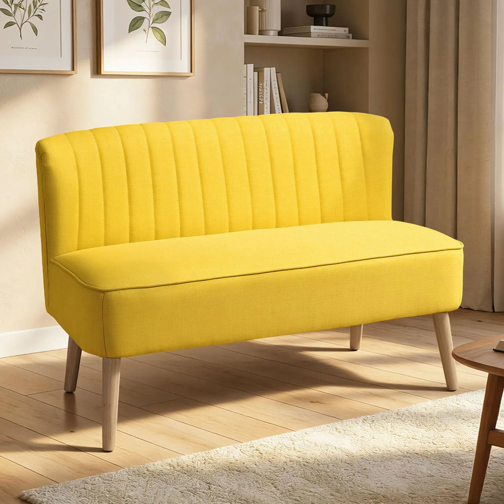 Geltona dvivietė sofa jaukioje svetainėje šalia knygų lentynos