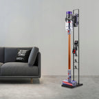 Dyson dulkių siurblys su priedais ant juodo metalo stovo svetainėje šalia pilkos sofos