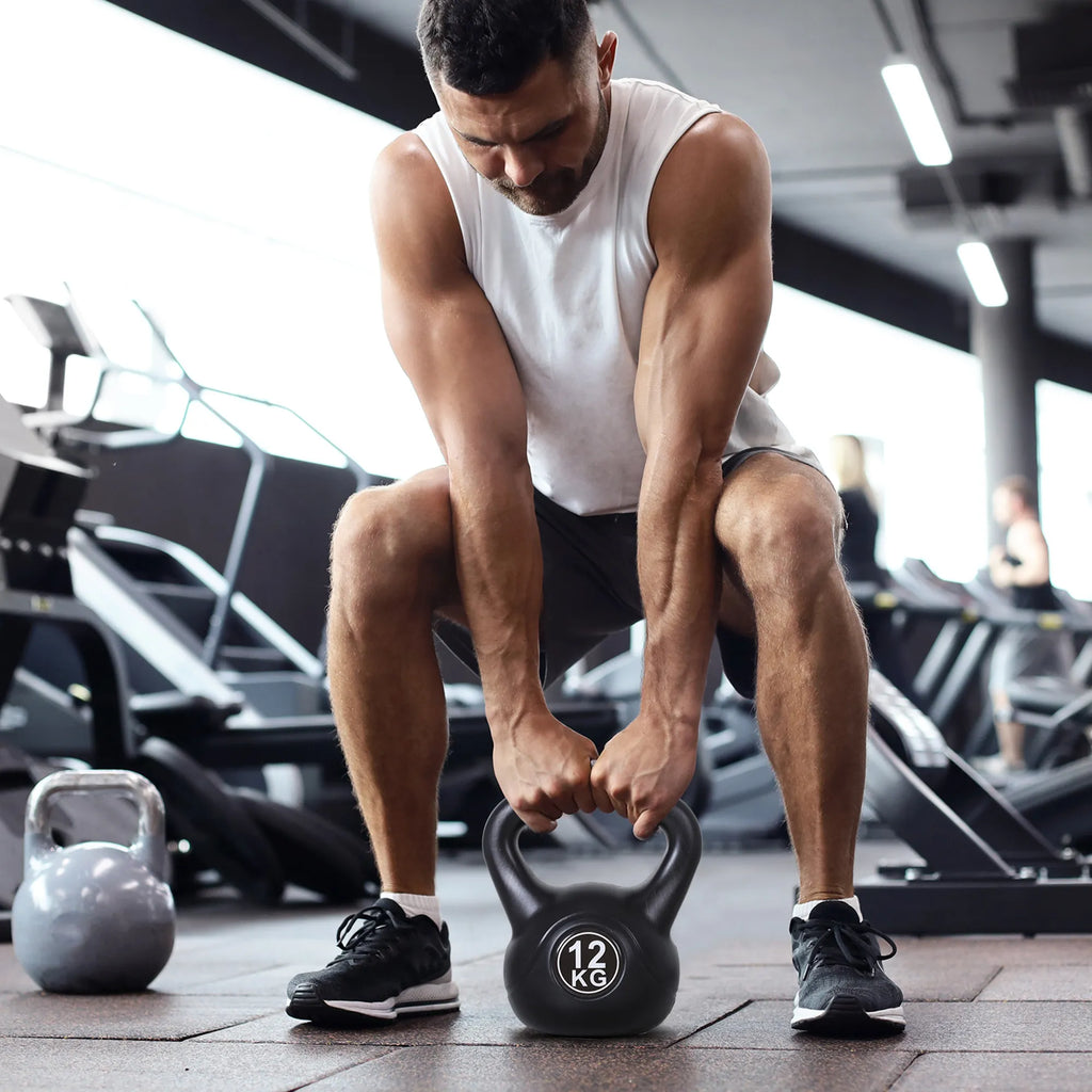 Vyras sporto salėje pakelia juodą kettlebell nuo grindų