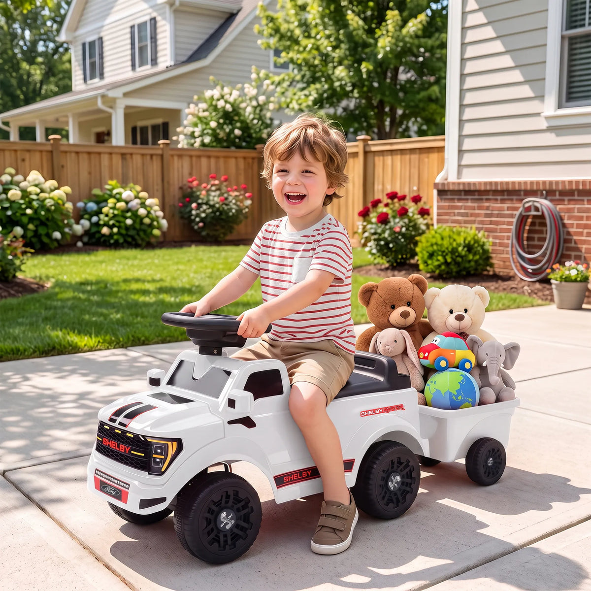 Berniukas linksmai sėdi automobiliuke su žaislų priekaba kieme